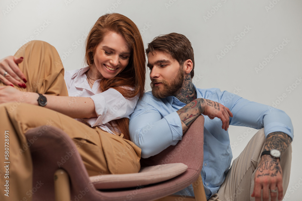 Happy beautiful young fashion couple are sitting in the studio. Beautiful red-haired young woman with a cute smile and a fashionable handsome man with a tattoo and a beard Happy beautiful young fashion couple are sitting in the studio. Beautiful red-haired young woman with a cute smile and a fashionable handsome man with a tattoo and a beard