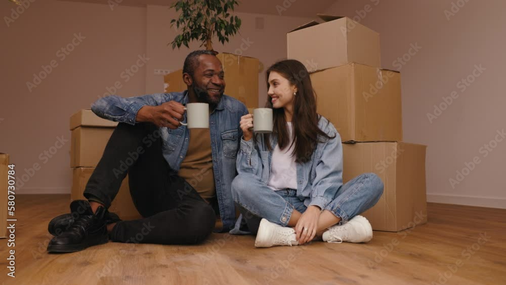 Happy african american man and caucasian woman moving to new home. Newly married mixed couple moving to new home and drinking coffee on the floor near cardboard boxes. Happy african american man and caucasian woman moving to new home. Newly married mixed couple moving to new home and drinking coffee on the floor near cardboard boxes.
