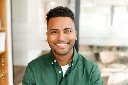 Canvas Print Portrait of happy satisfied Indian young businessman standing in the office and looking at the camera with light smile