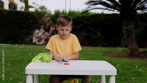 Child with autism has math therapy lesson: uses colorful Numicon shapes, counts, writes answers on paper; sits at white table in sunny garden with bright green grass