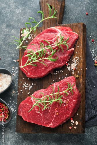 Raw beef steak. Marbled raw fresh Ribeye steak with rosemary, salt and pepper on cutting board on dark concrete background. Raw beef steak and spices for cooking.