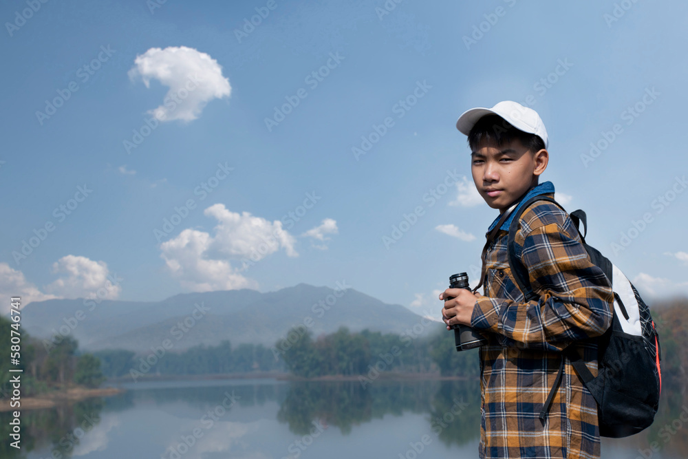 Asian boy in plaid shirt wears cap and has a backpack, holding a binoculars, standing on ridge reservoir in local national park to observe birds on tree branches and on sky and to watch fish in lake.