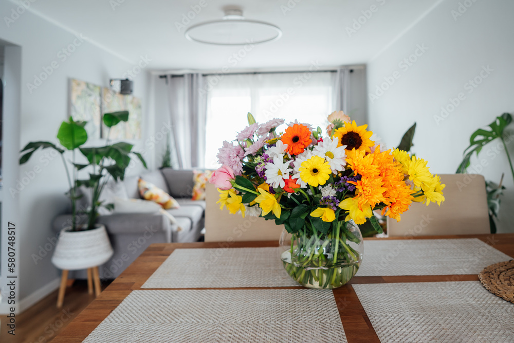 Kitchen counter table with focus on vase with huge multicolor various flower bouquet with blurred background of modern cozy living room with couch and green plants. Open space home interior design.