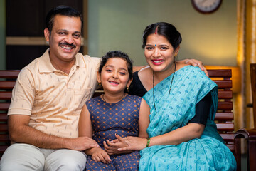 Happy smiling couple with daughter looking camera while sitting on sofa at home - concept of relationship, family photo posing and togetherness