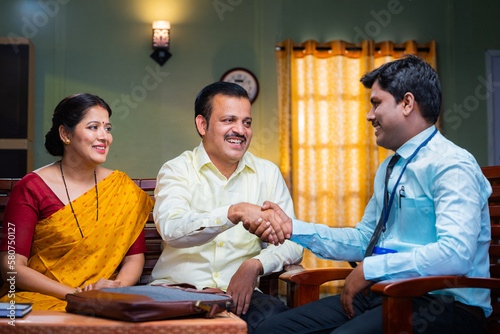 Banking officer greeting by shaking hand to couple by explaining about insurance at home - concept of financial adviser, doorstep service or support and investment planning