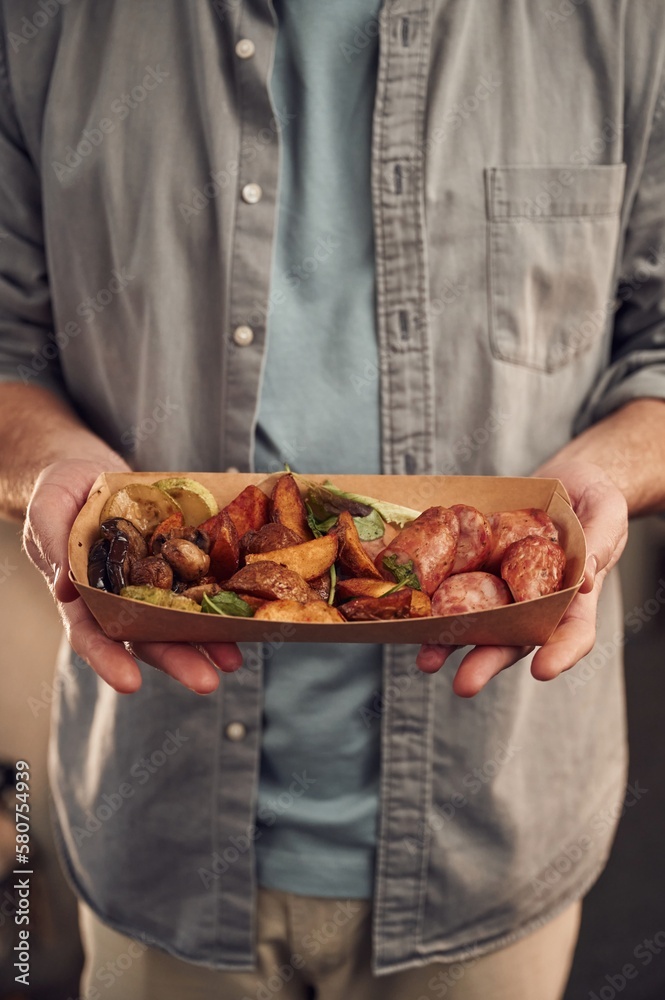 Close up view. Man in casual clothes is holding food that are in the paper eco box. Potatoes, sausages, mushrooms, zucchini