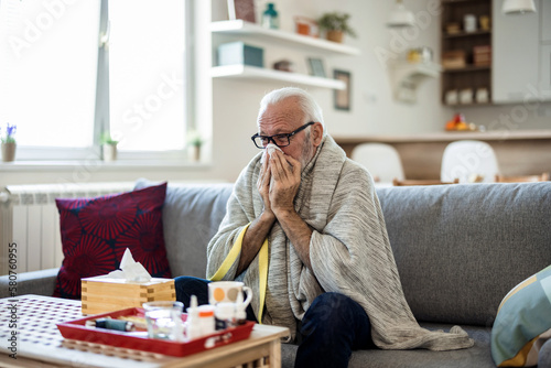 Senior man suffering from flu while sitting wrapped in a blanket on the sofa at home. Elderly Man blowing his nose while lying sick in bed at home. Man with a cold lying in sofa holding tissues photo