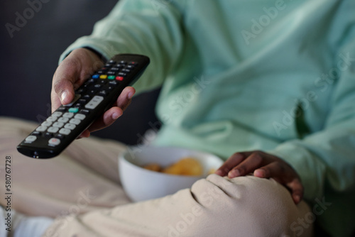 Close-up of little boy in casualwear pressing buttons on remote control while sitting on couch, having snack and choosing curious tv channel