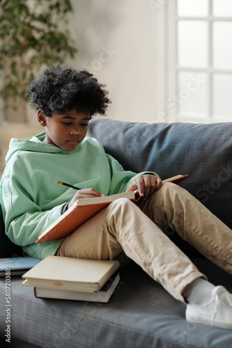 Serious African American schoolboy with open encyclopaedia on his knees reading article and making notes in copybook while preparing for lesson