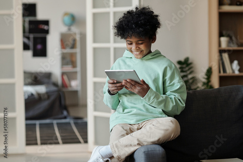 Smiling schoolboy in hoodie and pants watching online video while sitting on edge of couch in living room and looking at tablet screen