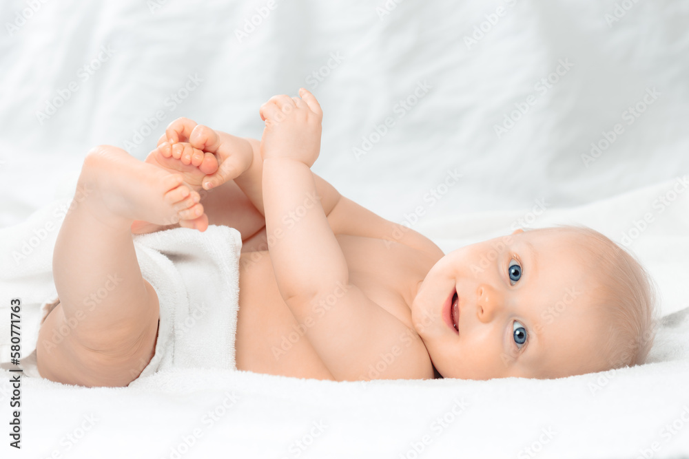 Cute happy baby with blue eyes lying on white bed and holding legs. Little smiling boy looking at camera. Child playing with his feet. Cute happy baby with blue eyes lying on white bed and holding legs. Little smiling boy looking at camera. Child playing with his feet.