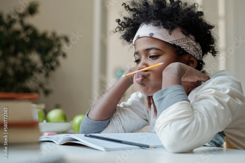 Distracted and lazy schoolboy reluctant to do homework sitting by desk with open copybook and holding pencil between nose and upper lip photo