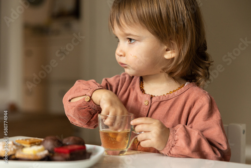 Little girl eats at the table and sticks her fingers into a glass of drink.