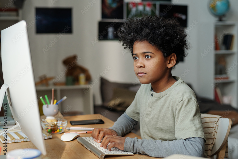 Serious schoolboy sitting in front of computer screen in living room and typing on keyboard while carrying out home assignment