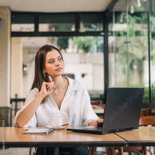 Vertical portrait developing a new business strategy. Young Caucasian business woman in formal clothes thinking about something and looking at laptop while sitting in modern office