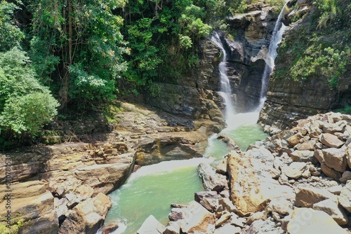 The spectacular waterfall Air Terjun Tengkulese on Flores which flows into a water basin surrounded by sunlit rainforest, rocks in the foreground. photo