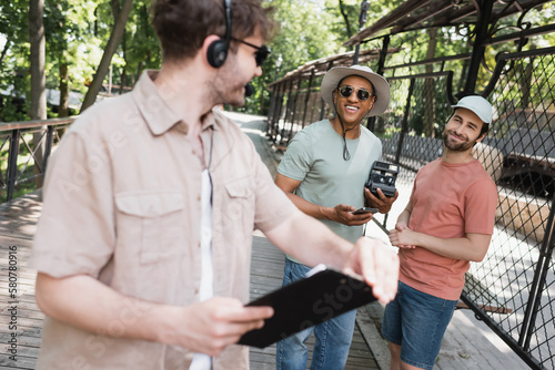 carefree multiethnic men with vintage camera and smartphone looking at blurred tour guide with clipboard in summer park. photo