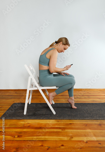 A young blonde girl in green sportswear is sitting on a white chair using her phone after doing yoga exercise. The concept of sports and correct posture for girls.