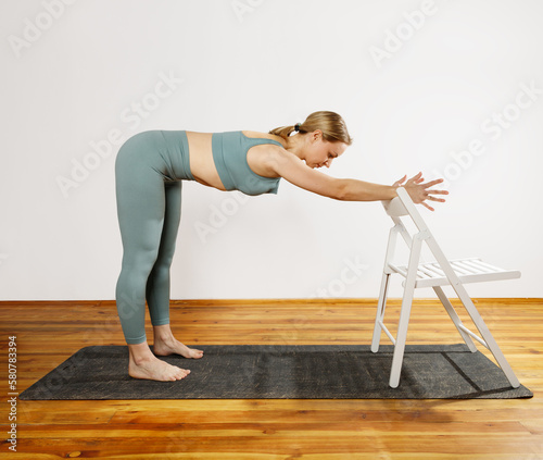 European blonde woman doing yoga in green sportswear practices sports pose indoors with a chair. Young slim woman in sportswear doing yoga for back