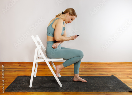A young blonde girl in green sportswear is sitting on a white chair using her phone after doing yoga exercise. The concept of sports and correct posture for girls.