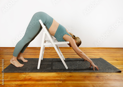 European blonde woman doing yoga in green sportswear practices sports pose indoors with a chair. Young slim woman in sportswear doing yoga for back