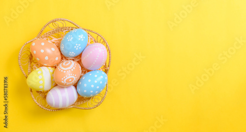 Happy easter celebration holiday. colourful pastel painted eggs in wicker basket nest decoration on a yellow background. Seasonal greeting gift card concept. Top view, flat lay, copy space.