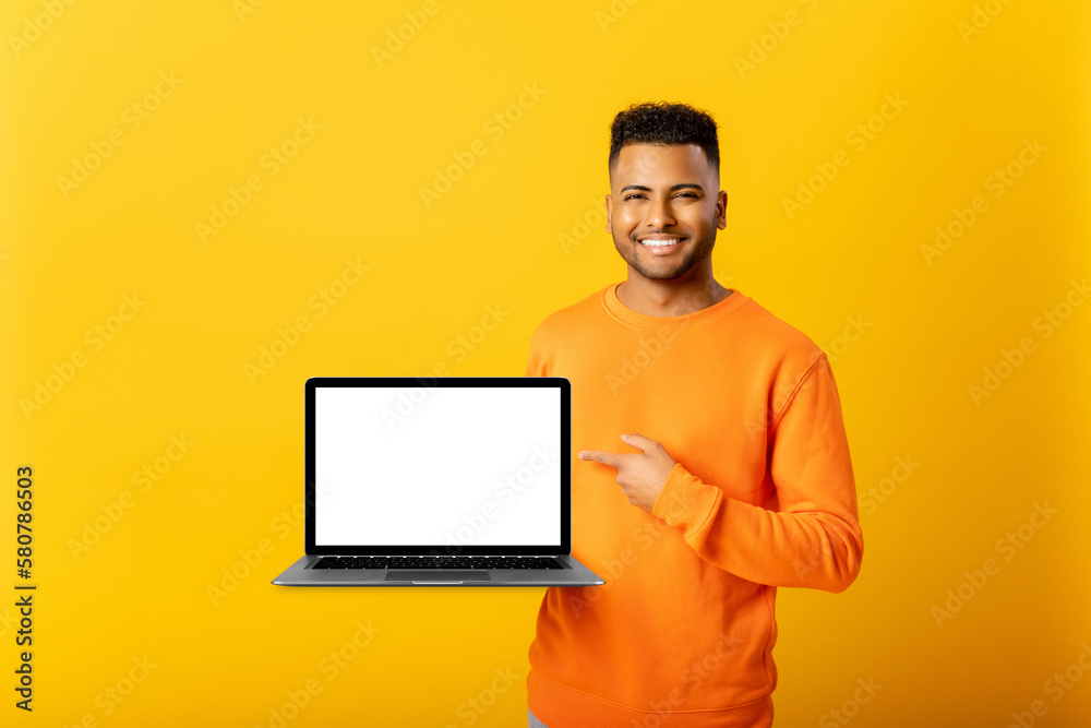 Positive optimistic indian guy freelancer or student showing laptop with empty screen and points finger on it, standing isolated on yellow, arab man presenting new app, mock-up, copy space