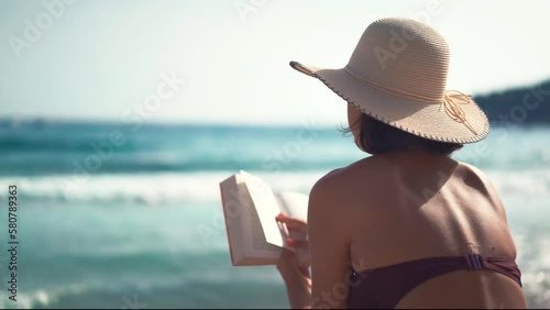 Beautiful young female with hat reading book on sandy beach enjoying her holiday next to sea. What book to read on vacation? Reading books on beach.