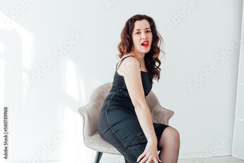 a brunette woman in a black dress sits on a chair in a bright room