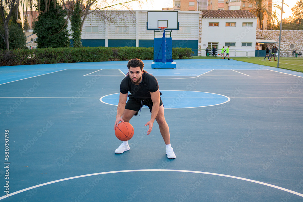 Fototapeta premium Afro Latin man playing basketball, bouncing the ball, on a court at sunset