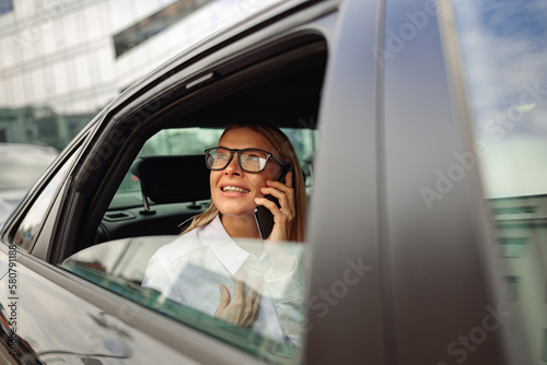 Businesswoman in eyeglasses traveling on back seat of car and talking phone while looking at window 