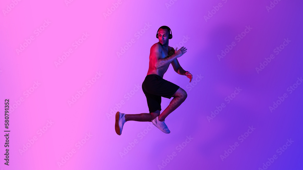 Athletic African American Male Jumping Wearing Headphones Over Purple Background