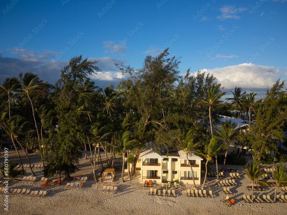 Hotel among palm trees on the beach. Beach chairs are placed on the shore. There are white clouds in the bright blue sky. Resort place, paradise, romance, vacation, rest.