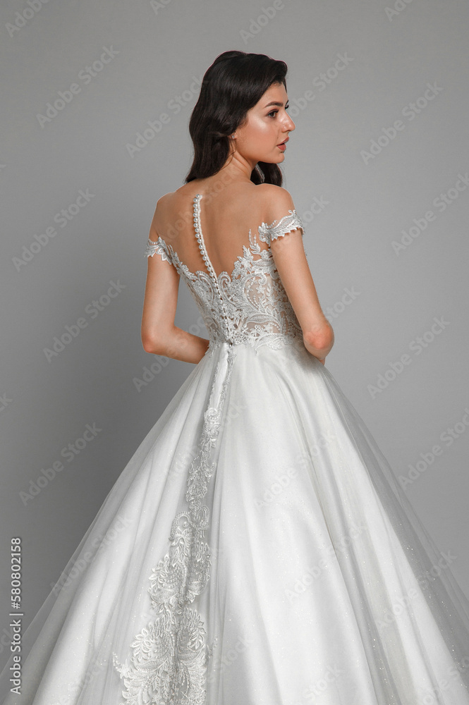 Close up of the back view portrait of young beautiful woman wearing white wedding dress with pearls. Elegant bride standing and posing in the studio. Close up of the back view portrait of young beautiful woman wearing white wedding dress with pearls. Elegant bride standing and posing in the studio.