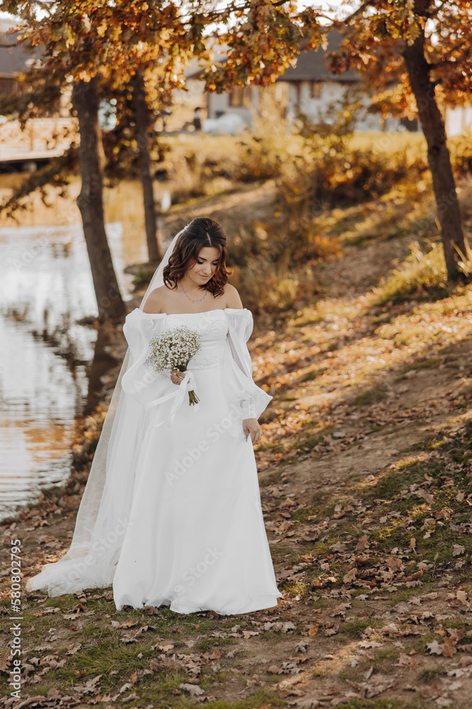 Awesome bride in a long wedding dress with a beautiful bouquet in hand and with charming smile. Under the tree in the beautiful sunset light in the park.