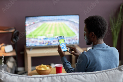 Canvas Print Back view of man holding smartphone with sports bets app on screen while watchin