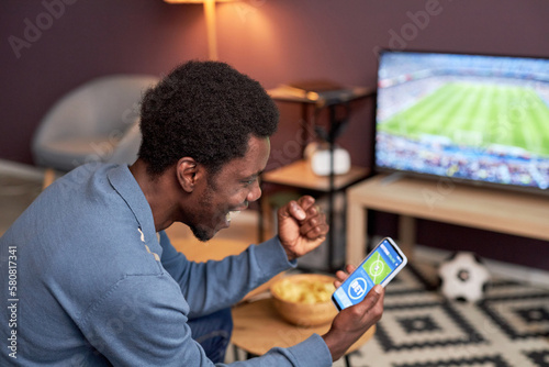 Fototapeta Naklejka Na Ścianę i Meble -  Side view portrait of black man holding smartphone with online sport bets app and cheering for football team