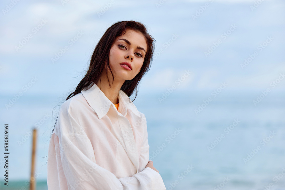 Portrait of a beautiful pensive woman with tanned skin in a white beach shirt with wet hair after swimming on the ocean beach sunset light with clouds, the concept of freedom and mental health