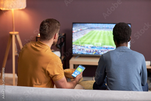 Back view of two men watching football match on TV and holding smartphone with online bets