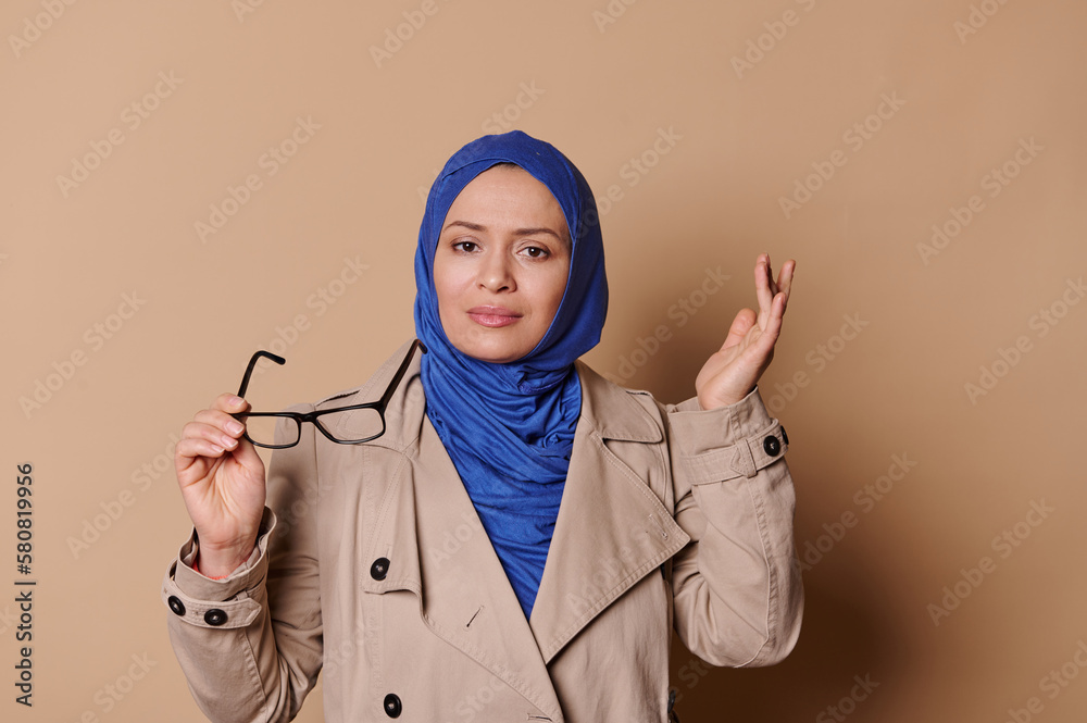 Doubtful, hesitant Middle-Eastern Muslim woman in blue hijab and casual classical coat, looking at camera, posing with stylish eyeglasses over isolated beige background. Optics. Style. Arab fashion Doubtful, hesitant Middle-Eastern Muslim woman in blue hijab and casual classical coat, looking at camera, posing with stylish eyeglasses over isolated beige background. Optics. Style. Arab fashion