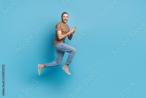 Full length photo of funny cute man wear beige t-shirt jumping high texting modern device emtpy space isolated blue color background