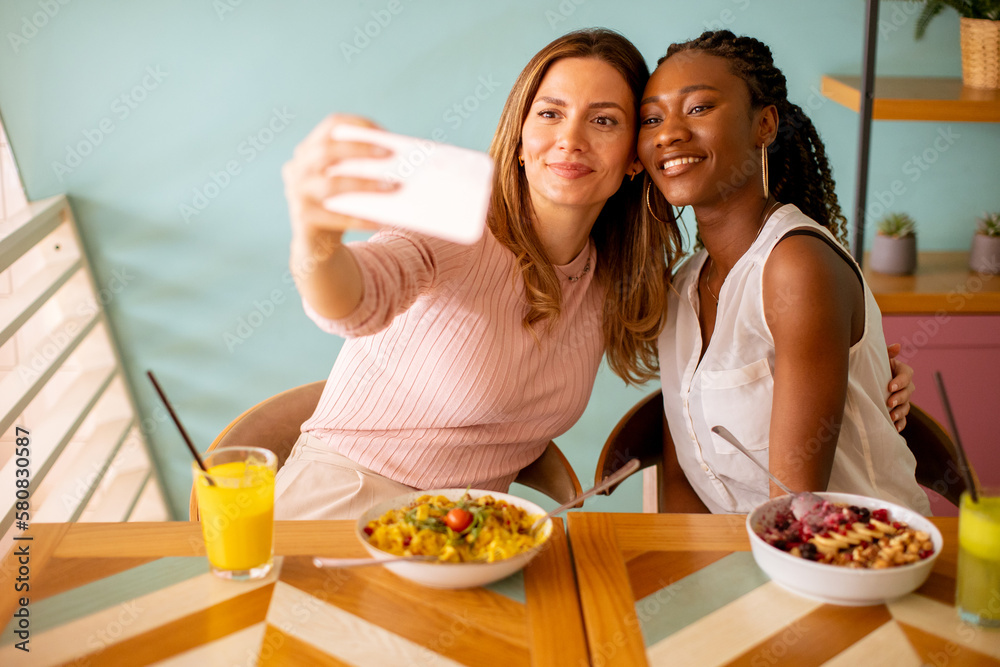 Two young women, caucasian and black one, taking selfie with mobile phone in the cafe