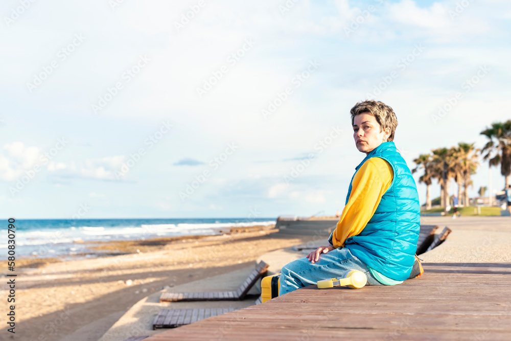 Sad Woman in a blue jacket with a yellow suitcase and headphone on the seaside on hot sunny day arrived in a tourist town.  Travel  Lifestyle concept