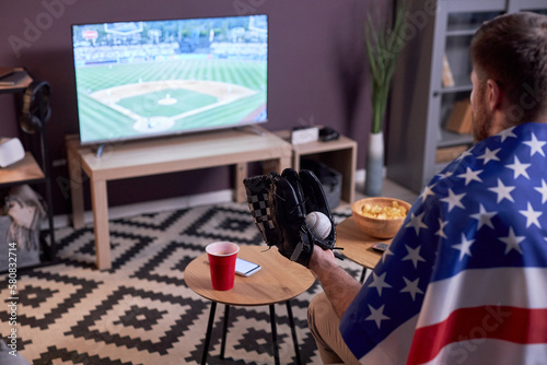 Wall Mural Side view of sports fan watching baseball match at home on TV and wearing USA fl