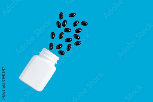 White bottle with black therapeutic capsules, vitamin pills or drugs for treatment on blue background, medicine and healthcare concept, top view