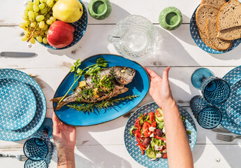 Gilt-head bream also known as Orata grilled on a barbeque grill served by a woman on a white wooden table with vegetable salad and fruits. Healthy seafood preparation concept photo.