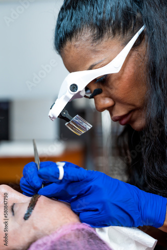 Side view of crop black female microblading master in magnification goggles and gloves applying permanent makeup on eyebrows in professional beauty salon