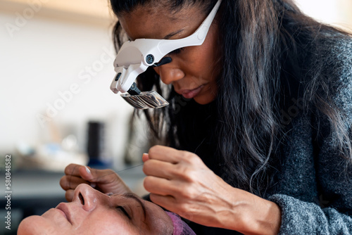 Crop black cosmetologist in magnification goggles doing facial hair removal threading procedure to client in beauty salon