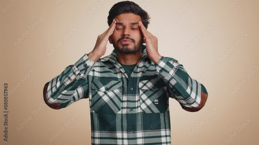 Handsome indian bearded man in shirt rubbing temples to cure headache problem, suffering from tension, migaine, stress, grimacing in pain, high blood pressure isolated alone on beige wall background Handsome indian bearded man in shirt rubbing temples to cure headache problem, suffering from tension, migaine, stress, grimacing in pain, high blood pressure isolated alone on beige wall background