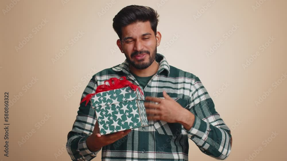 Positive smiling young indian man presenting birthday gift box stretches out hands, offer wrapped present career bonus, celebrating party, sale. Bearded hindu guy isolated on beige studio background Positive smiling young indian man presenting birthday gift box stretches out hands, offer wrapped present career bonus, celebrating party, sale. Bearded hindu guy isolated on beige studio background